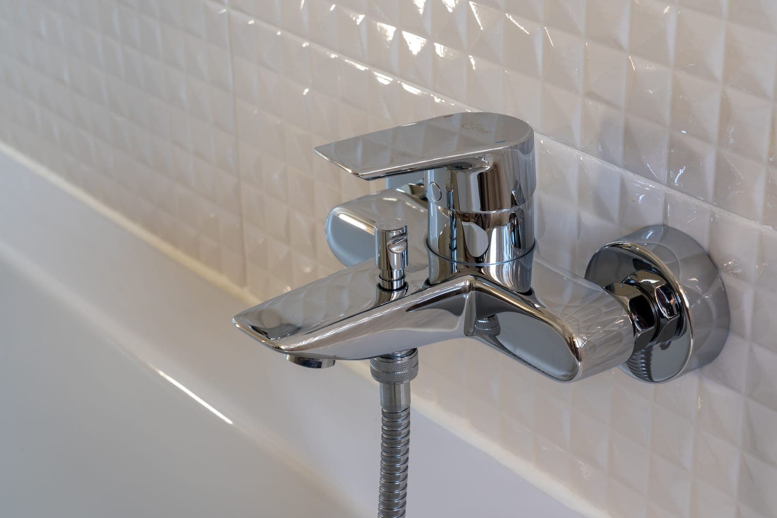 A chrome bath and shower mixer on white tiled wall, with the shower hose connected below it