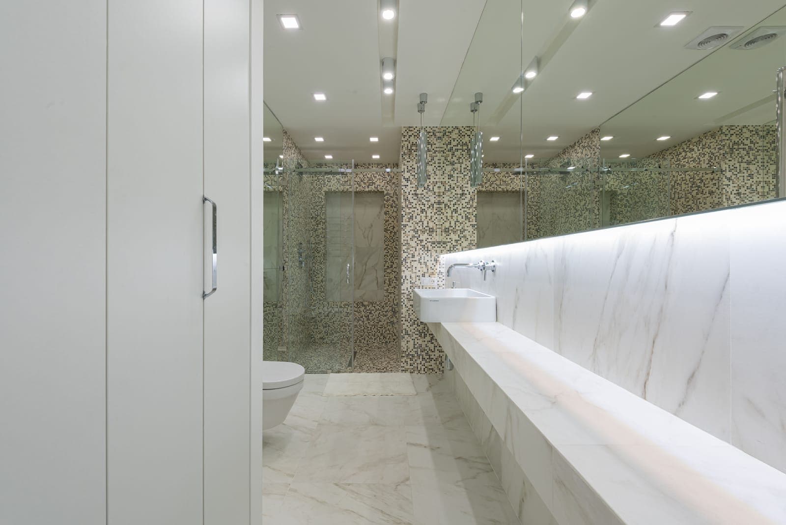 A windowless marble bathroom with recessed ceiling lights and ceiling vents above the vanity