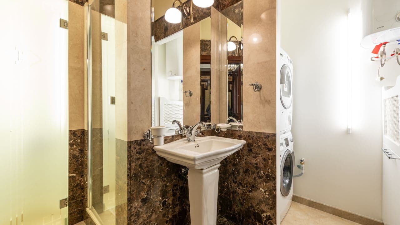 A rental bathroom with a pedestal sink, mirror, shower enclosure and a stacked washer-dryer