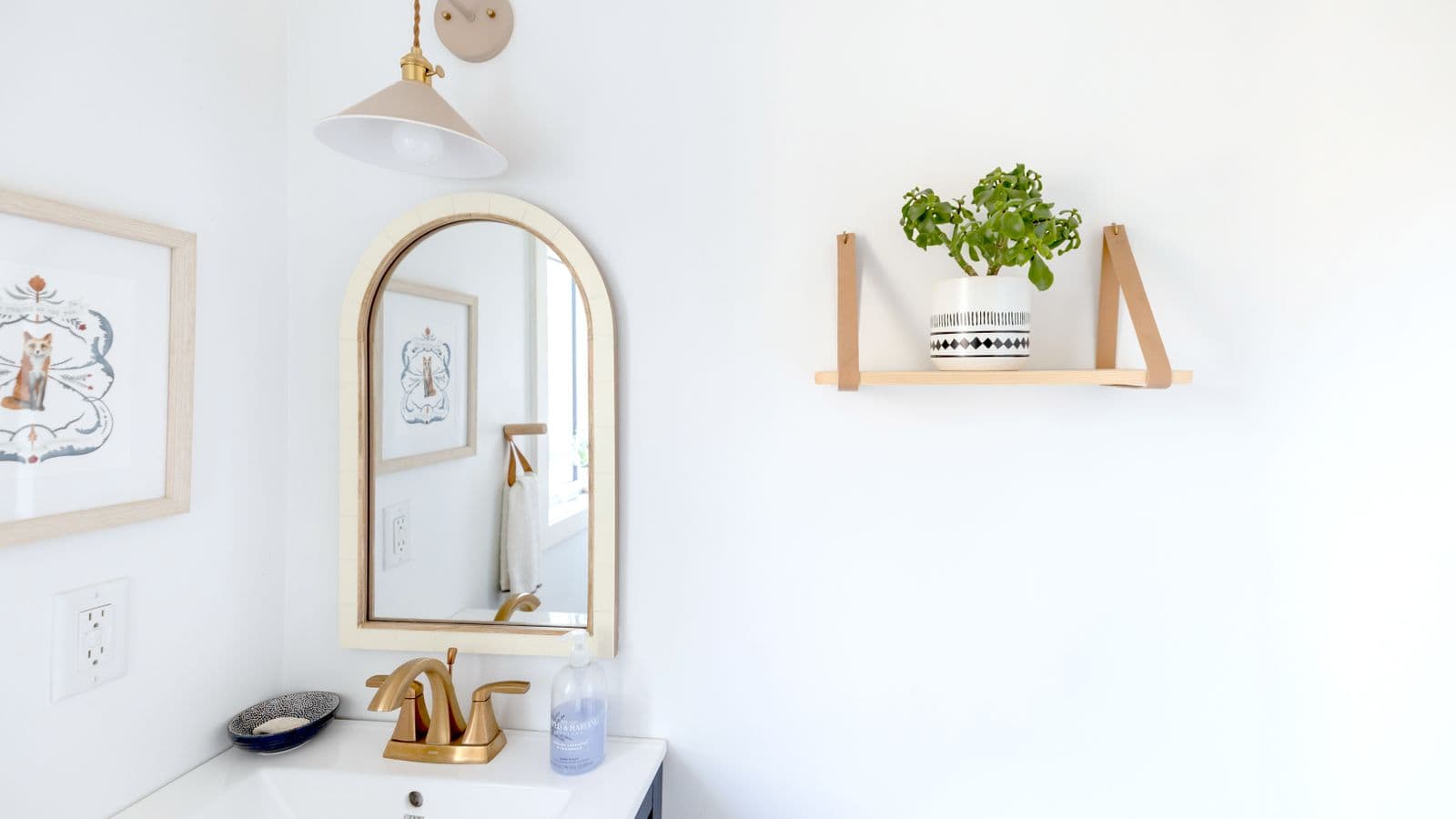 A small bright bathroom with an arched mirror, gold faucet and a wooden wall shelf