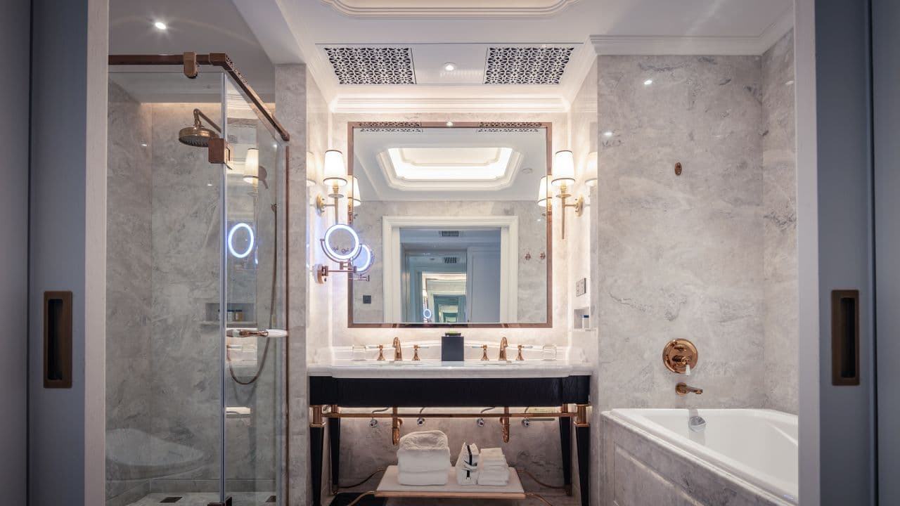 A marble hotel bathroom with a glass shower, lit mirror and folded white towels on a shelf