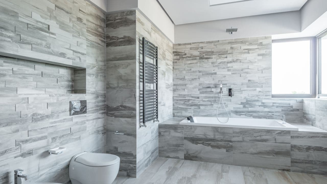 A gray stone-tiled bathroom with a tiled floor, a built-in bathtub and a wall-mounted towel rail