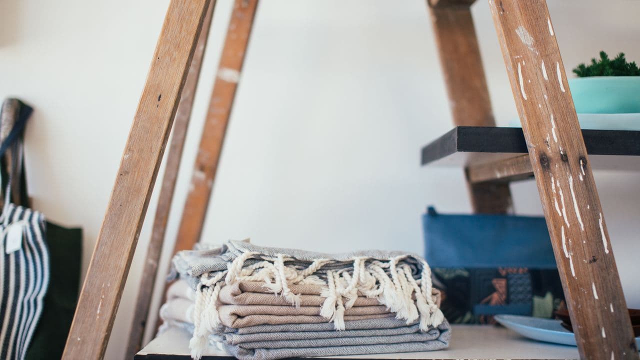 A folded stack of fringed cotton towels on a wooden shelf beside a step ladder