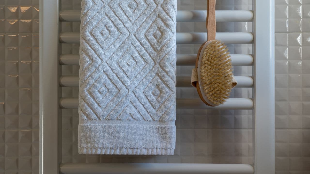 A white patterned towel and a wooden body brush hanging on a white heated towel rail