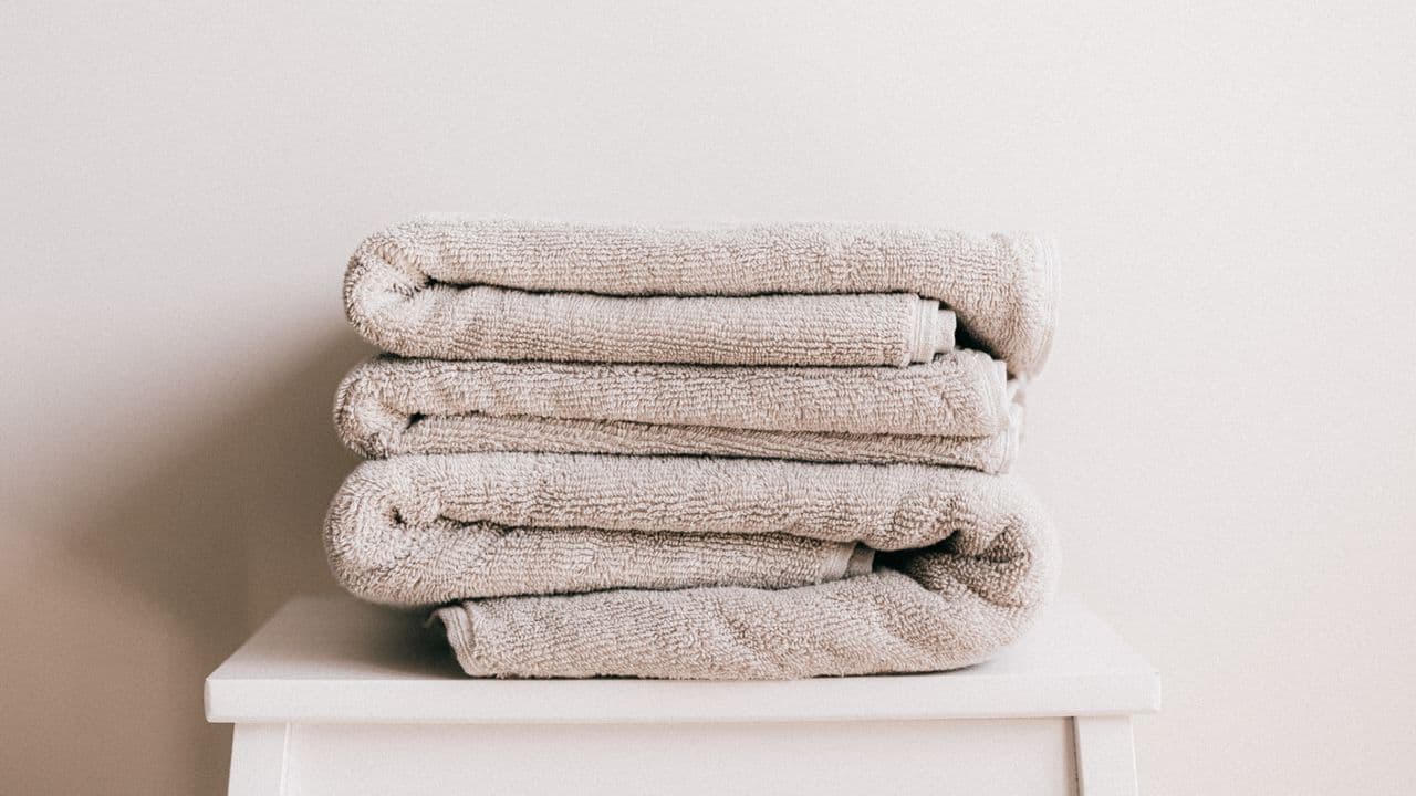 A stack of folded beige bath towels on a white stool against a pale wall