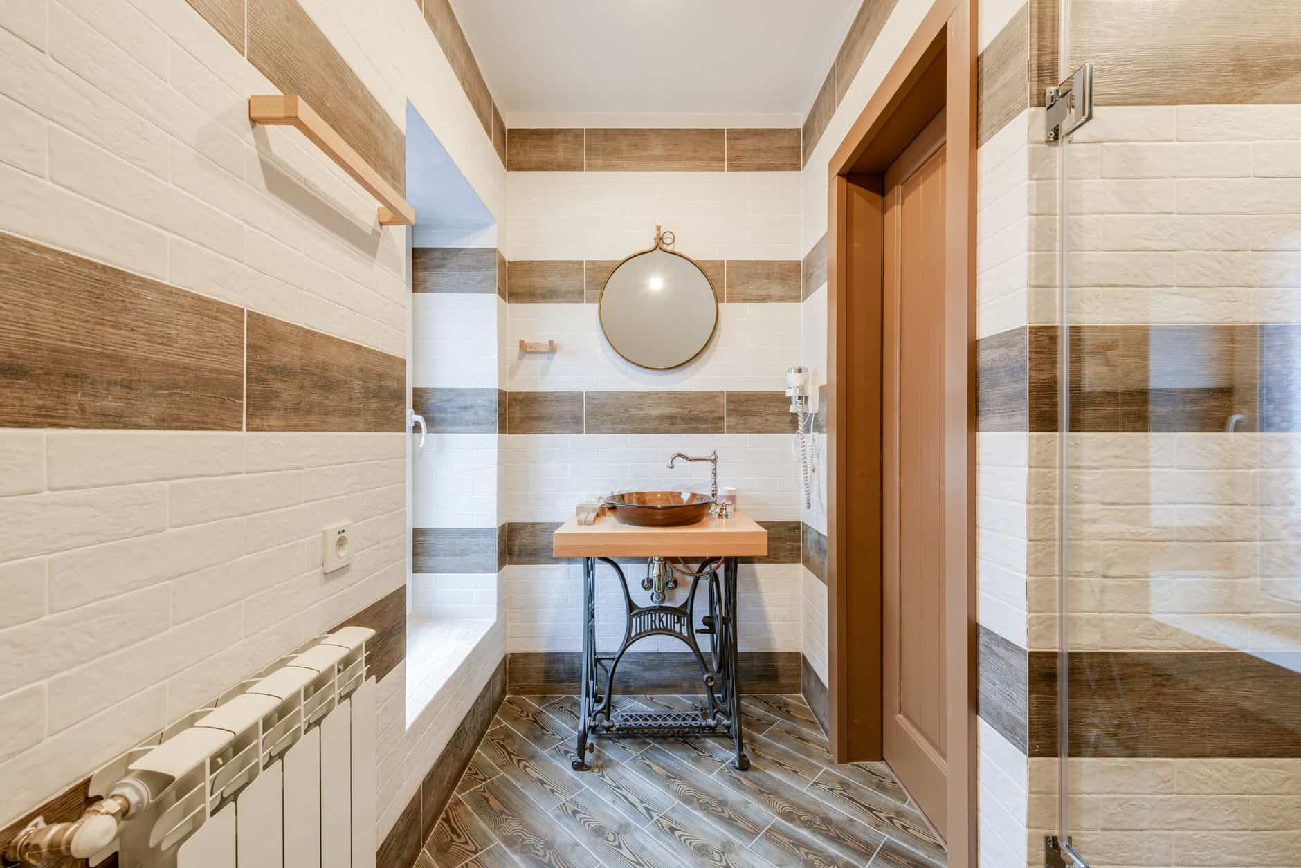 A white panel radiator on the wall of a tiled bathroom, beside a basin and round mirror
